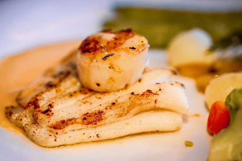 Un gros plan d'un plat gastronomique magnifiquement présenté, peut-être une noix de Saint-Jacques poêlée avec une sauce vive, accompagnée d'un verre de vin blanc